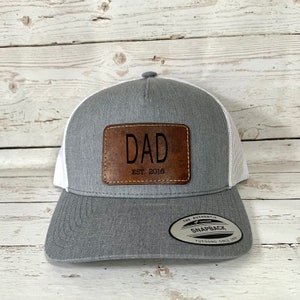 Peut inclure: Casquette de camionneur grise et blanche avec un patch en cuir marron qui dit "DAD EST. 2016".