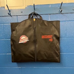 Peut inclure: Un sac de vêtement noir avec une fermeture éclair. Le sac a un logo rouge et blanc qui dit "Oak Ridge Aeros" et un texte rouge qui dit "Greenway #19".