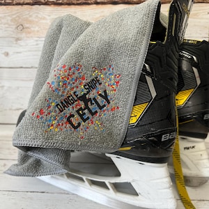 Peut inclure: Serviette en microfibre grise avec un motif coloré qui dit "Dangle Snipe Celly". La serviette est drapé sur une paire de patins de hockey sur glace noirs et blancs.