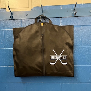 Peut inclure: Un sac de vêtement noir avec une fermeture éclair. Le sac a un graphique blanc de deux bâtons de hockey croisés avec le texte "GREENWAY #19" imprimé dessus.