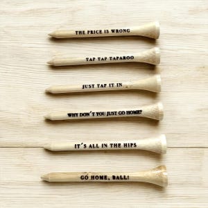Peut inclure: Six tees de golf en bois avec des phrases humoristiques imprimées en noir. Les phrases incluent "THE PRICE IS WRONG", "TAP TAP TAPAROO", "JUST TAP IT IN", "WHY DON'T YOU JUST GO HOME?", "IT'S ALL IN THE HIPS" et "GO HOME, BALL!".