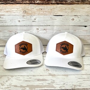Peut inclure: Deux casquettes de camionneur blanches avec des patchs en cuir marron. Les patchs sont hexagonaux et présentent le texte "Shooter McGavin" et une silhouette noire d'une main pointant une arme à feu.