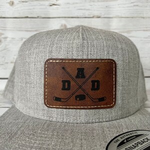 May include: Grey and brown baseball cap with a leather patch featuring the letters "DAD" with a hockey stick design and the letter "A" in the centre.