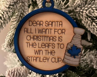 May include: A wooden Christmas ornament with a blue border and a hockey stick and puck design. The ornament reads "Dear Santa all I want for Christmas is the Leafs to win the Stanley Cup."