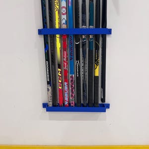 Peut inclure: Un support bleu pour crosses de hockey fixé à un mur blanc, contenant plusieurs crosses. Les crosses présentent diverses couleurs et marques, dont "CCM" et "Bauer". Le support est placé au-dessus d'une bande jaune.