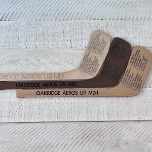 Peut inclure: Trois crosses de hockey en bois avec texte gravé, dont "OAKRIDGE AEROS U9 MD1" et les noms des joueurs. Les crosses sont de différentes nuances de marron et de beige, sur fond de bois blanc.