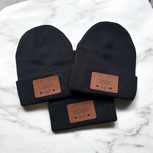 May include: Three black knit beanies with brown leather patches. The patches have hockey stick graphics and personalised names and numbers: Moon #88, Jones #4, and Dickson #97.