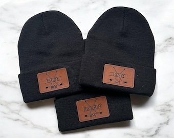 May include: Three black knit beanies with brown leather patches. The patches have hockey stick graphics and personalized names and numbers: Moon #88, Jones #4, and Dickson #97.