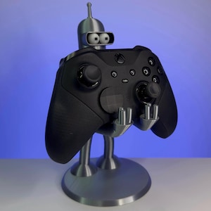 May include: A silver 3D printed robot holding a black Xbox controller. The robot has a round head with two large eyes and a small antenna. The controller has a textured black surface and a silver button.