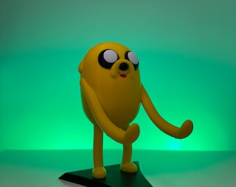 May include: A yellow Jake the Dog figurine from Adventure Time, with black eyes and a small red tongue. The figure is standing on a black triangular base. The background is a gradient of green.