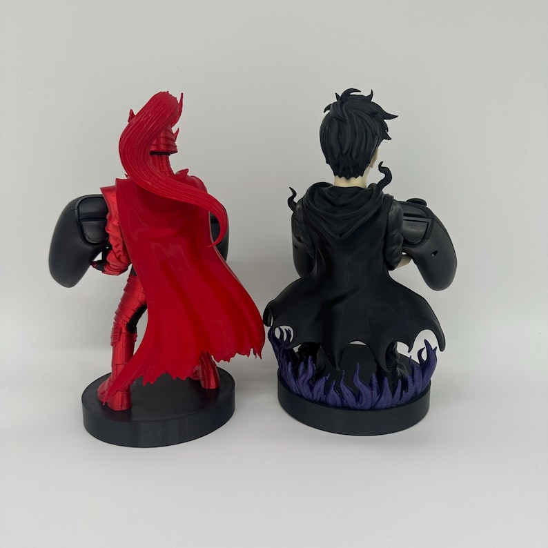 May include: Two detailed figurines, one in red armor with a flowing cape and the other in a black cloak, each holding a controller. The figurines are on black circular bases. The second figurine's base has purple flame details.