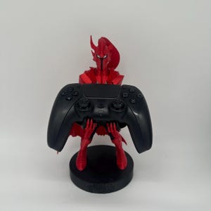 May include: A red, 3D-printed warrior figure holding a black video game controller. The figure is on a black circular base. The controller has buttons and directional controls.
