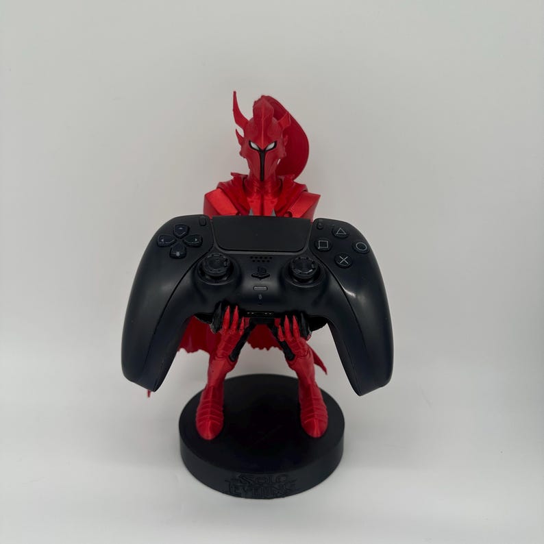 May include: A red, 3D-printed warrior figure holding a black video game controller. The figure is on a black circular base. The controller has buttons and directional controls.
