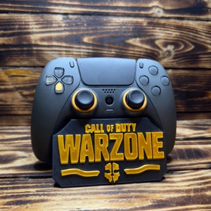 Puede incluir: Mando de juegos negro y dorado con un soporte con el logo "Call of Duty Warzone". El mando tiene detalles dorados. El soporte es negro con letras doradas, sobre un fondo de madera.
