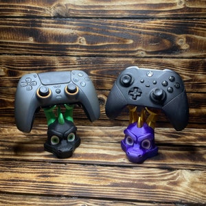 May include: Two video game controllers are displayed on dragon-shaped stands. The controller on the left is gray and has a black dragon stand. The controller on the right is black and has a purple dragon stand.