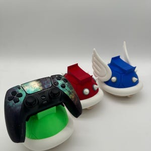 May include: A black video game controller with a galaxy design sits on a green shell-shaped stand. Two shell-shaped console accessories in red and blue, with white accents and spikes. The blue shell has white wings.