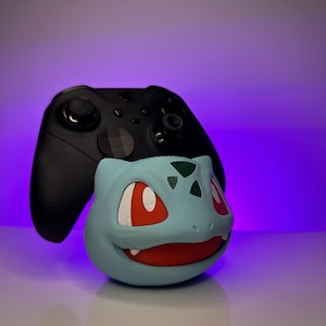 May include: A blue and green Pokemon character figurine with red eyes and a toothy grin. The figurine is holding a black Xbox controller.