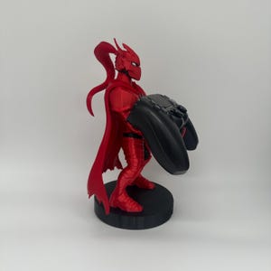 May include: A red figurine of a character in a suit of armor, holding a black video game controller. The figurine has a cape and is standing on a black circular base. The character's helmet has a long, flowing ponytail.