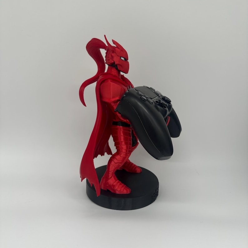 May include: A red figurine of a character in a suit of armor, holding a black video game controller. The figurine has a cape and is standing on a black circular base. The character's helmet has a long, flowing ponytail.