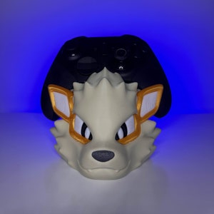 Peut inclure: Une tête de loup imprimée en 3D de couleur crème, avec des accents dorés et blancs, conçue pour maintenir une manette de jeu noire. La manette repose sur la tête du loup, créant un présentoir unique pour les accessoires de jeu.