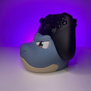 May include: A blue and beige 3D printed character with a black Xbox controller resting on top. The controller is facing the camera and the character is facing to the right.