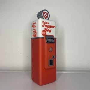 May include: A red and white vintage-style vending machine with the text "Drink Jugger-Nog 10&cent;" on the front. The machine has a coin slot and a button.