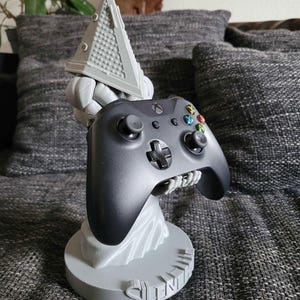 Può includere: Un supporto per controller di videogiochi stampato in 3D di colore grigio che presenta Pyramid Head della serie di videogiochi Silent Hill. Il supporto tiene un controller Xbox nero.