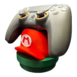May include: A gray gaming controller rests on a red Mario hat-shaped stand with a white "M" emblem. The stand is placed on a green, cylindrical base. The controller has orange accents.