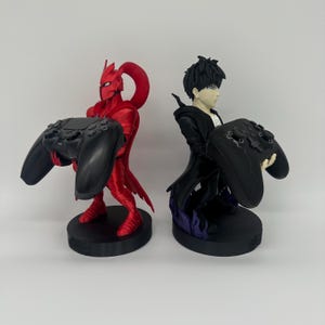 May include: Two gaming controller stands. One is a red figure with a helmet and cape holding a black controller. The other is a black figure with a white shirt holding a black controller. Both figures are on black bases.