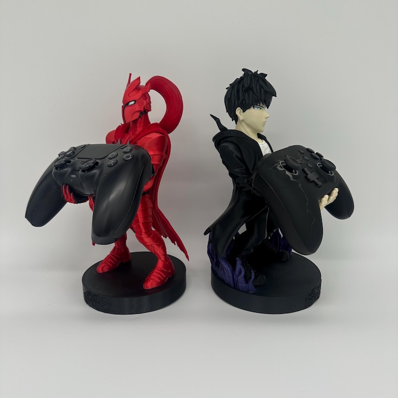 May include: Two gaming controller stands. One is a red figure with a helmet and cape holding a black controller. The other is a black figure with a white shirt holding a black controller. Both figures are on black bases.