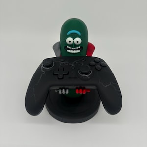 May include: A black video game controller with lightning bolt designs, resting on a black stand. A green pickle character with a red cape is behind the controller. The controller has gray and red accents.