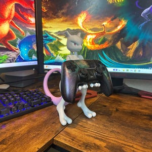 May include: A grey Mewtwo figure supporting a black games controller with a galaxy design. The figure is supported by white legs and a pink tail, resting on a wooden desk. The background shows computer monitors displaying colourful fantasy art.