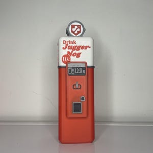 May include: A red and white vintage-style vending machine with the text "Drink Jugger-Nog 10&cent;" on the front. The machine has a small red cross on the top and a slot for coins.