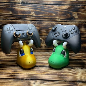 Op de afbeelding: Twee videogamecontrollers, een zwart en een grijs, zijn gemonteerd op een standaard. De standaarden zijn gevormd als cartoonfiguren, een groen en een geel.