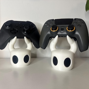 Pode incluir: Dois controladores de videogame pretos são exibidos em suportes brancos em forma de cabeça de um personagem do videogame Hollow Knight. Os suportes são brancos com olhos pretos.