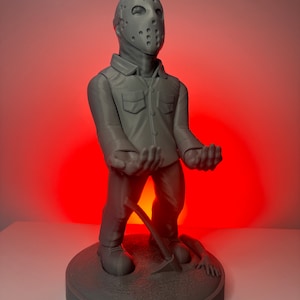 May include: A gray 3D printed figurine of a horror movie character wearing a hockey mask and holding a machete. The figurine is standing on a base with a hand reaching out from the ground.