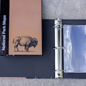 May include: A black binder with a clear plastic pocket on the right side. The front cover of the binder has a brown background with the text "National Park Maps" and a black and white illustration of a buffalo.