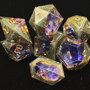 May include: A set of seven polyhedral dice for tabletop games. The dice are clear with a teal and gold border, and feature a purple and blue galaxy-like interior. The numbers are gold. The dice are arranged on a black surface.
