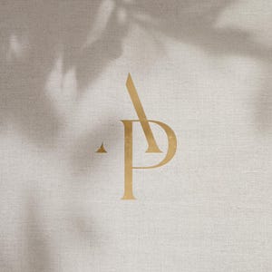 May include: Gold monogram logo featuring the letters "A" and "P" intertwined, set against a neutral, textured background. The design includes a small triangle and a leaf shadow, creating a minimalist and elegant aesthetic.