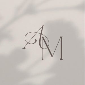 May include: A minimalist design featuring the intertwined letters "A" and "M" in a soft, muted brown hue. The elegant, serif font is set against a neutral, off-white background with subtle shadow effects.