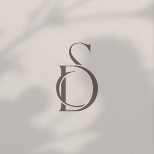 Könnte beinhalten: Ein minimalistisches Logo-Design mit den ineinander verschlungenen Buchstaben "S" und "D" in einer weichen, gedämpften Brauntönung. Die Buchstaben sind elegant mit einer Serifenschrift gestaltet, vor einem neutralen Hintergrund mit subtilen Schatteneffekten.