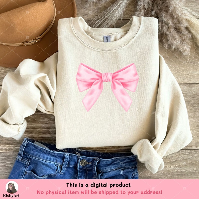 May include: A beige sweatshirt with a pink satin bow on the front. The sweatshirt is laying on a wooden surface with a brown hat and blue jeans.