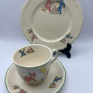 Könnte beinhalten: Ein Vintage-Kinder-Tee-Set mit weißem Hintergrund und grünem Rand. Die Teetasse und die Untertasse zeigen ein Cartoon-Bild eines Hasen in einem rosa Kleid. Der größere Teller zeigt ein Cartoon-Bild von zwei Hasen, einer in einem blauen Anzug und einer in einem rosa Kleid.