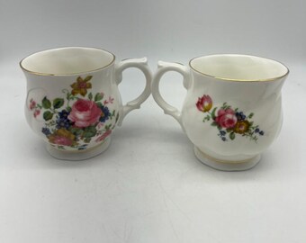 May include: Two white ceramic mugs with floral designs. The mugs have gold trim around the rim and a floral pattern with pink, yellow, and blue flowers.