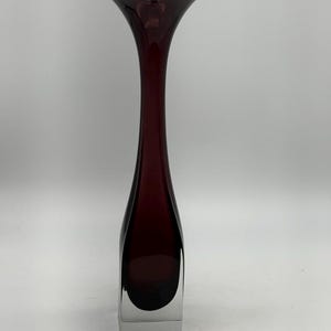 Peut inclure: Un vase en verre haut et élancé doté d'un corps couleur prune profonde qui se transforme en une base carrée solide et transparente. Le col du vase se rétrécit avant de s'évaser vers le haut en un rebord large, organique et asymétrique.