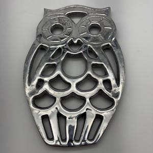 Peut inclure: Un dessous de plat argenté en forme de hibou. Le dessous de plat présente un motif ajouré détaillé, avec des formes découpées formant les traits du hibou. Le métal a un aspect légèrement texturé.