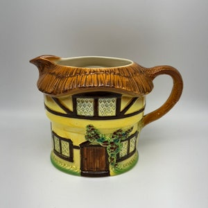 May include: A yellow ceramic pitcher shaped like a cottage with a thatched roof, brown door, and green vines.