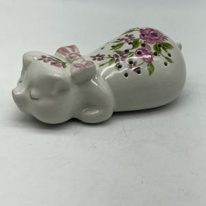 Może przedstawiać: Biała ceramiczna figurka świnki z różowymi i fioletowymi detalami kwiatowymi. Świnka leży na boku z zamkniętymi oczami. Figurka ma kokardkę na głowie i jest ozdobiona małymi otworami.