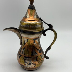 May include: An ornate metal coffee pot with a long spout and a curved handle. The pot features intricate designs in copper, silver, and gold tones, with a detailed lid and a decorative finial. The body of the pot has a black background with figures.