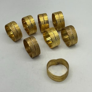 Puede incluir: Ocho anillos de servilleta dorados con un acabado texturizado y una banda elevada. Los anillos están dispuestos en círculo sobre una superficie blanca. Hay un anillo adicional en el centro de la imagen.